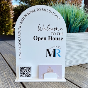 May include: White open house sign with a black and blue logo that says "MR". The sign also says "Have a look around and prepare to fall in love! Welcome to the Open House". There is a QR code and a business card holder on the sign.