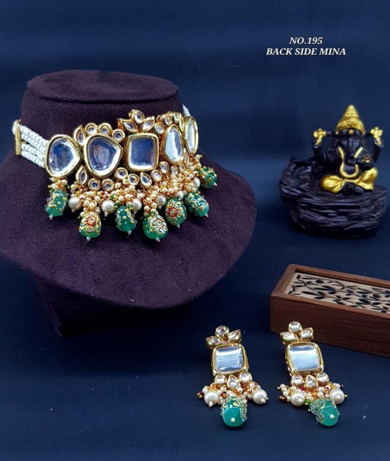 May include: A gold necklace with a pearl and green bead design. The necklace features a large, square, mirrored pendant with smaller mirrored accents. The necklace is paired with matching earrings with a similar design. The image text reads "NO. 195 BACK SIDE MINA".