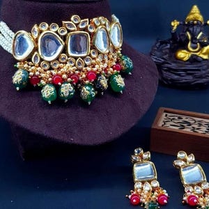 May include: A gold necklace with a white pearl strand and a large, ornate pendant featuring mirrored glass stones. The pendant is surrounded by smaller, gold-toned stones and has a dangling design with green and red beads. Matching earrings with a similar design are also included.