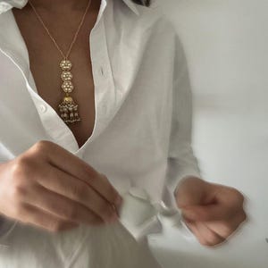 May include: A woman wearing a white button-down shirt with a long gold necklace featuring a pendant with multiple circular designs and a tassel.