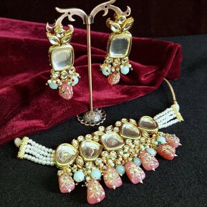 May include: A gold and white choker necklace with pink and blue beads and a large, ornate pendant. The necklace is on a red velvet background.