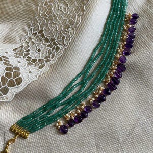 Peut inclure: Un collier à plusieurs rangs avec des perles vert émeraude et des accents d'améthyste. Le collier a des fermoirs dorés et de petites perles blanches. Le bijou est présenté sur une surface blanche texturée.