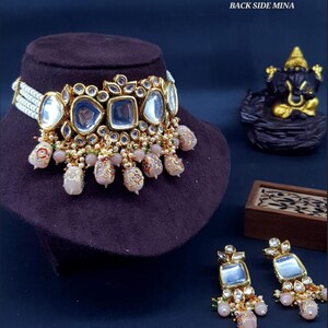 May include: A gold choker necklace with a mirrored design and pink and white beads. Matching earrings with a mirrored design and pink and white beads. The necklace and earrings are labeled "NO.195 BACK SIDE MINA".