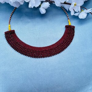 Op de afbeelding: Een dieprode kralenketting met een gebogen ontwerp, tegen een lichtblauwe achtergrond. Witte bloemen zijn zichtbaar in het bovenste deel van de afbeelding. De ketting is een choker-stijl, geschikt voor formele of informele gelegenheden.