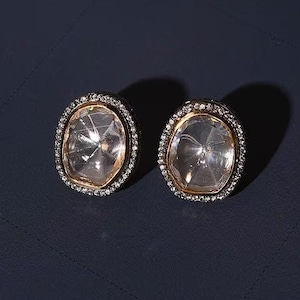 May include: A pair of oval-shaped earrings with clear crystal stones set in gold-toned metal frames. The frames are accented with a row of small, clear stones.