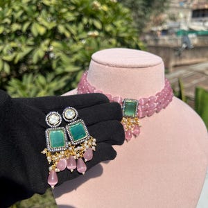 May include: A pink beaded choker necklace with a green stone pendant and matching earrings. The earrings have a green stone surrounded by clear rhinestones and dangle with pink beads.