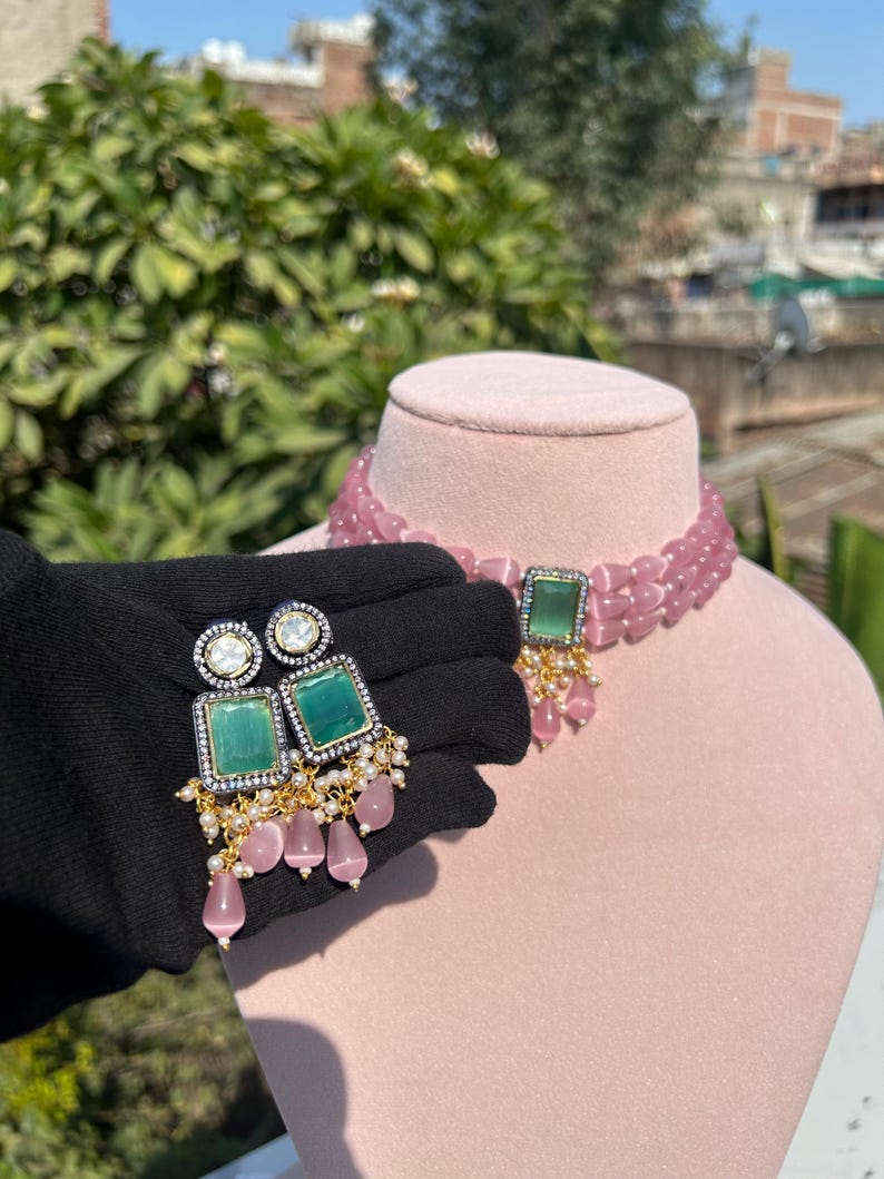 May include: A pink beaded choker necklace with a green stone pendant and matching earrings. The earrings have a green stone surrounded by clear rhinestones and dangle with pink beads.