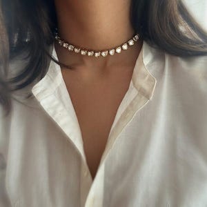 May include: A gold choker necklace with clear, faceted stones. The necklace is worn by a person with long, dark hair and a white shirt.