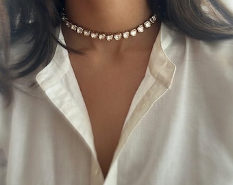 May include: A gold choker necklace with clear, faceted stones. The necklace is worn by a person with long, dark hair and a white shirt.
