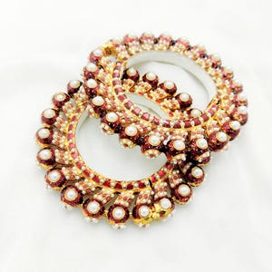 Peut inclure: Deux bracelets dorés ornés de détails rouges et blancs. Chaque bracelet est orné de petites perles blanches rondes et de bijoux rouges. Les bracelets sont présentés sur un fond blanc, mettant en valeur leur design et leur savoir-faire.