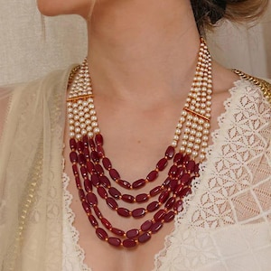 May include: A multi-strand necklace featuring red and white beads. The necklace is crafted from oval-shaped red beads and small white pearls. The necklace is layered and has a gold clasp.