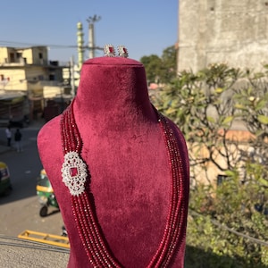 May include: A red beaded necklace with a silver pendant and matching earrings. The necklace is made of multiple strands of beads and has a decorative silver pendant with a red stone in the center. The earrings are small and have a similar design to the pendant.