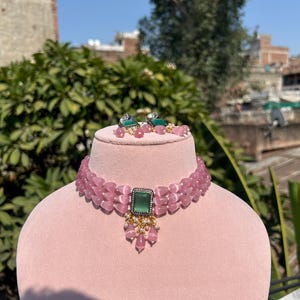 May include: A pink choker necklace with a green gemstone pendant. The necklace is made of multiple strands of pink beads and has a delicate design. The pendant is a square-shaped green gemstone with a gold setting.