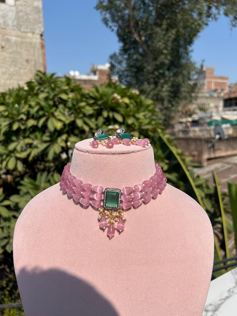 May include: A pink choker necklace with a green gemstone pendant. The necklace is made of multiple strands of pink beads and has a delicate design. The pendant is a square-shaped green gemstone with a gold setting.