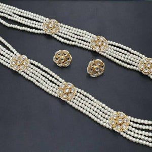 Peut inclure: Un collier de perles à cinq rangs avec des accents dorés et des boucles d'oreilles assorties. Chaque rang est orné d'une pierre ronde et transparente.