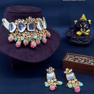 May include: A gold and white choker necklace with a large, clear, beveled glass centerpiece. The necklace is adorned with pink and green beads and pearls. The necklace is displayed on a black velvet surface. The image also shows a pair of matching earrings with a similar design.