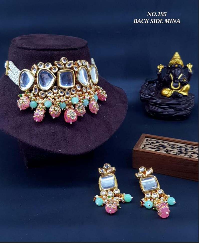 May include: A gold and white choker necklace with a large, clear, beveled glass centerpiece. The necklace is adorned with pink and green beads and pearls. The necklace is displayed on a black velvet surface. The image also shows a pair of matching earrings with a similar design.