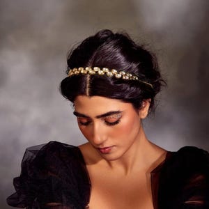May include: A woman with dark hair is wearing a gold headband with small round stones. She is wearing a black sheer top with puffed sleeves.