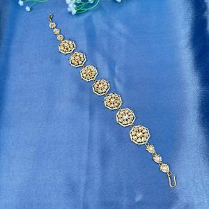 Może przedstawiać: Złoty nakrycie głowy z serią okrągłych, inspirowanych kwiatami wzorów, każdy ozdobiony małymi, przezroczystymi kamieniami szlachetnymi. Nakrycie głowy jest umieszczone na niebieskim tle z białymi akcentami kwiatowymi.