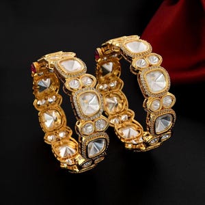 May include: Two gold bangles with intricate designs featuring large, clear, faceted stones. The bangles are set with smaller, round stones around the larger stones.
