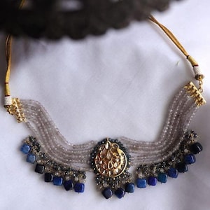 May include: A gold necklace with a large, ornate pendant and multiple strands of grey beads. The pendant is surrounded by smaller blue stones. The necklace is accented with blue square-shaped stones.