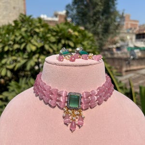 May include: A pink beaded choker necklace with a green gemstone pendant. The necklace has three rows of beads and the pendant is surrounded by smaller white beads.