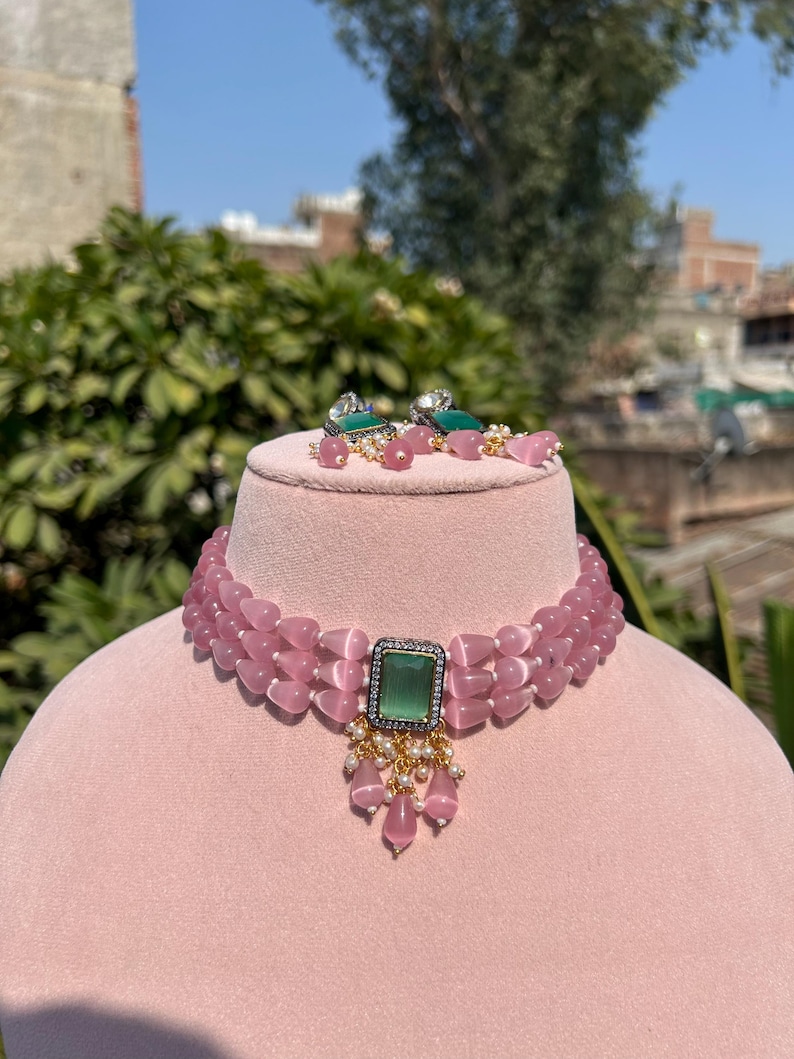 May include: A pink beaded choker necklace with a green gemstone pendant. The necklace has three rows of beads and the pendant is surrounded by smaller white beads.
