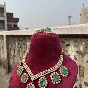 May include: A gold necklace with a V-shaped design and five emerald green stones. The necklace is on a red velvet display stand. There are also matching emerald green earrings on the stand.