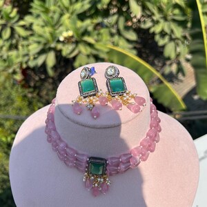 May include: A pink beaded choker necklace with a green gemstone pendant and matching earrings. The necklace has a double strand of beads and the pendant is surrounded by smaller white stones.