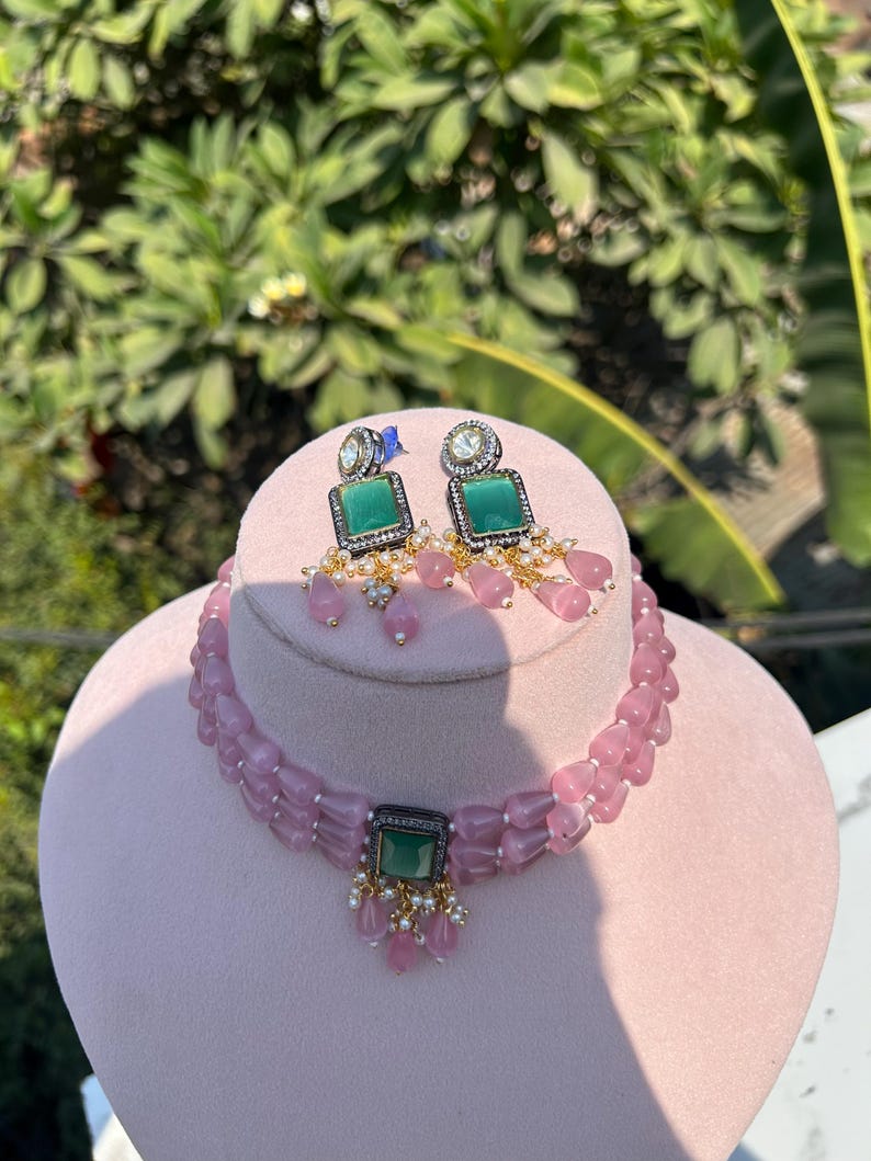 May include: A pink beaded choker necklace with a green gemstone pendant and matching earrings. The necklace has a double strand of beads and the pendant is surrounded by smaller white stones.
