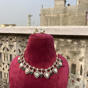 May include: A gold necklace with a white stone pendant and green accents. The necklace is on a red velvet display stand. The necklace is also available as earrings.