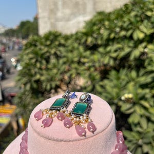 May include: A pink stone necklace with a green and silver pendant. The necklace is displayed on a pink stand with matching earrings featuring green stones and silver accents.
