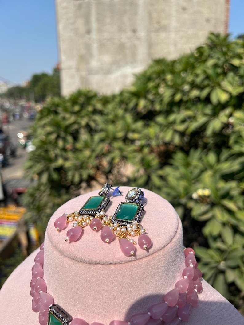 May include: A pink stone necklace with a green and silver pendant. The necklace is displayed on a pink stand with matching earrings featuring green stones and silver accents.