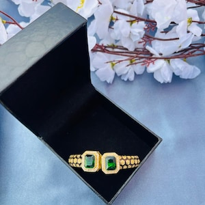 Op de afbeelding: Een goudkleurige armband met twee vierkante smaragdgroene edelstenen. De armband wordt gepresenteerd in een open zwarte doos, met witte bloemen op de achtergrond. De armband heeft een textuur.
