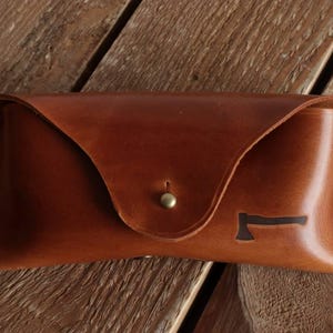 May include: A tan leather glasses case with a triangular flap closure secured by a brass stud. The case features a small, dark brown embossed axe icon on the lower right side and is constructed with visible stitching along the side edges.