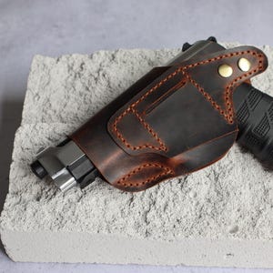 Op de afbeelding: Een donkerbruine lederen holster met zichtbare oranje stiksels en twee messingkleurige metalen drukknopen. De holster is gevormd voor een handvuurwapen, waarvan de metalen loop en de zwarte, getextureerde greep gedeeltelijk zichtbaar zijn.
