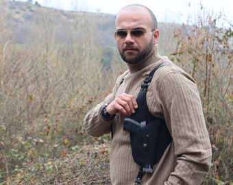 May include: A person wearing a brown turtleneck sweater and sunglasses is holding a black leather holster with a gun inside. The holster is worn across the chest and has a strap that goes over the shoulder.