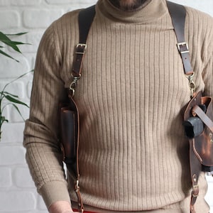 Puede incluir: Una correa de cámara de cuero marrón con dos bolsas y correas ajustables. La correa se lleva sobre los hombros de una persona.