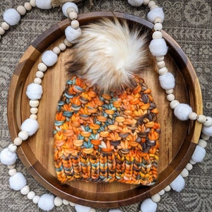 May include: A colorful knitted beanie with a fluffy pom pom. The beanie is made with a variety of orange, brown, and blue yarns. The beanie is sitting on a wooden tray with a white beaded garland.