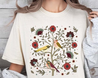 May include: A cream-colored t-shirt with a colorful floral design featuring birds and leaves. The design is centered on the shirt and is surrounded by a white border.