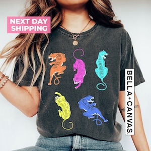 Puede incluir: Una camiseta gris oscuro con un gr&aacute;fico colorido de cinco tigres. Los tigres est&aacute;n en varias poses y est&aacute;n delineados en negro. El texto "BELLA + CANVAS" est&aacute; impreso en el lado derecho de la camiseta.