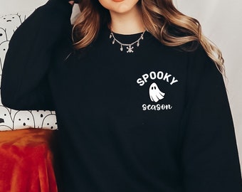 May include: A woman wearing a black witch hat and a black sweatshirt with a white ghost and the text "Spooky Season". She is holding a white pillow with a black ghost pattern.