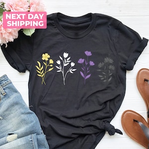 May include: Black t-shirt with a row of four stylized flowers in yellow, white, purple, and gray. The flowers are arranged in a line across the center of the shirt. The shirt is on a white wooden surface.