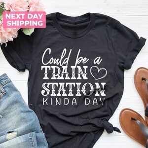 May include: Black t-shirt with white text that reads "Could be a Train Station Kinda Day" with a white heart symbol.