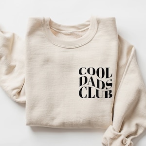 May include: A beige sweatshirt with the text "COOL DADS CLUB" printed in black on the front.