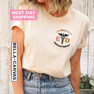 May include: A beige t-shirt with a black graphic of a medical symbol and the text "Emergency Department" in a circle with a pink, orange, and yellow color scheme.
