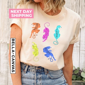 Puede incluir: Una camiseta beige con un estampado gr&aacute;fico de cinco tigres coloridos en diferentes poses. Los tigres est&aacute;n delineados en negro y rellenos de colores brillantes, incluyendo naranja, rosa, azul y amarillo.