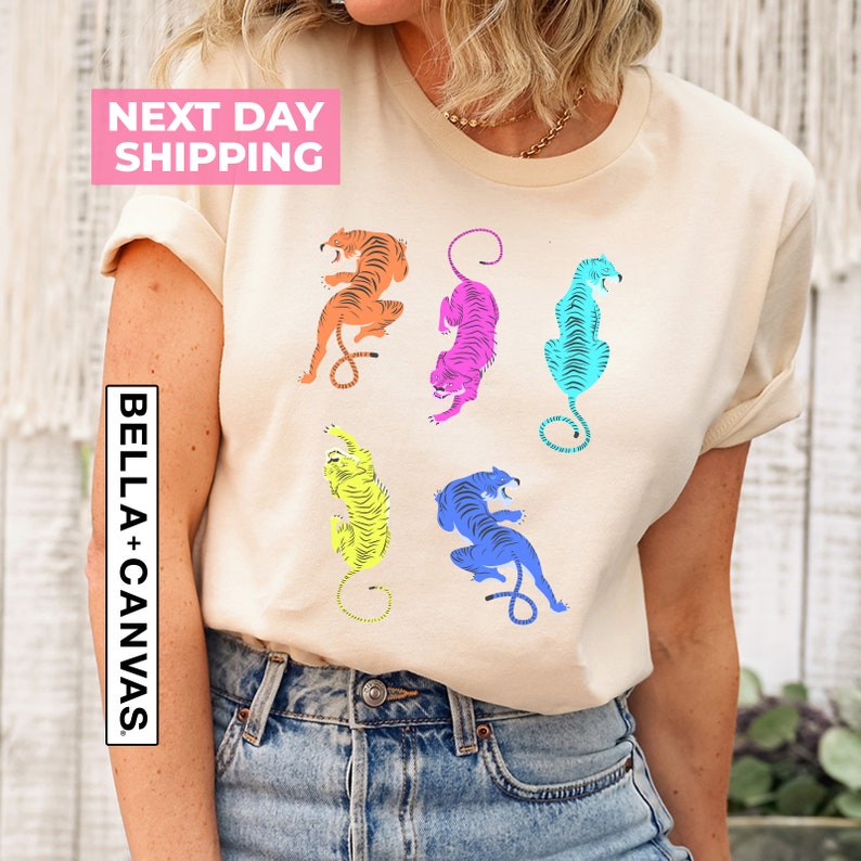 Puede incluir: Una camiseta beige con un estampado gr&aacute;fico de cinco tigres coloridos en diferentes poses. Los tigres est&aacute;n delineados en negro y rellenos de colores brillantes, incluyendo naranja, rosa, azul y amarillo.