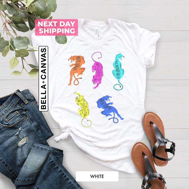 Puede incluir: Camiseta blanca con un gr&aacute;fico colorido de cinco tigres en diferentes poses. Los tigres est&aacute;n en tonos de naranja, amarillo, rosa, azul y turquesa.