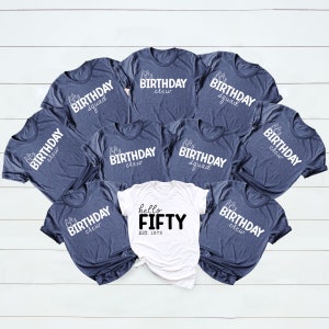 May include: A set of nine navy blue t-shirts with white text that reads "fifty birthday crew" and "fifty birthday squad". One white t-shirt with black text that reads "hello fifty est. 1973".